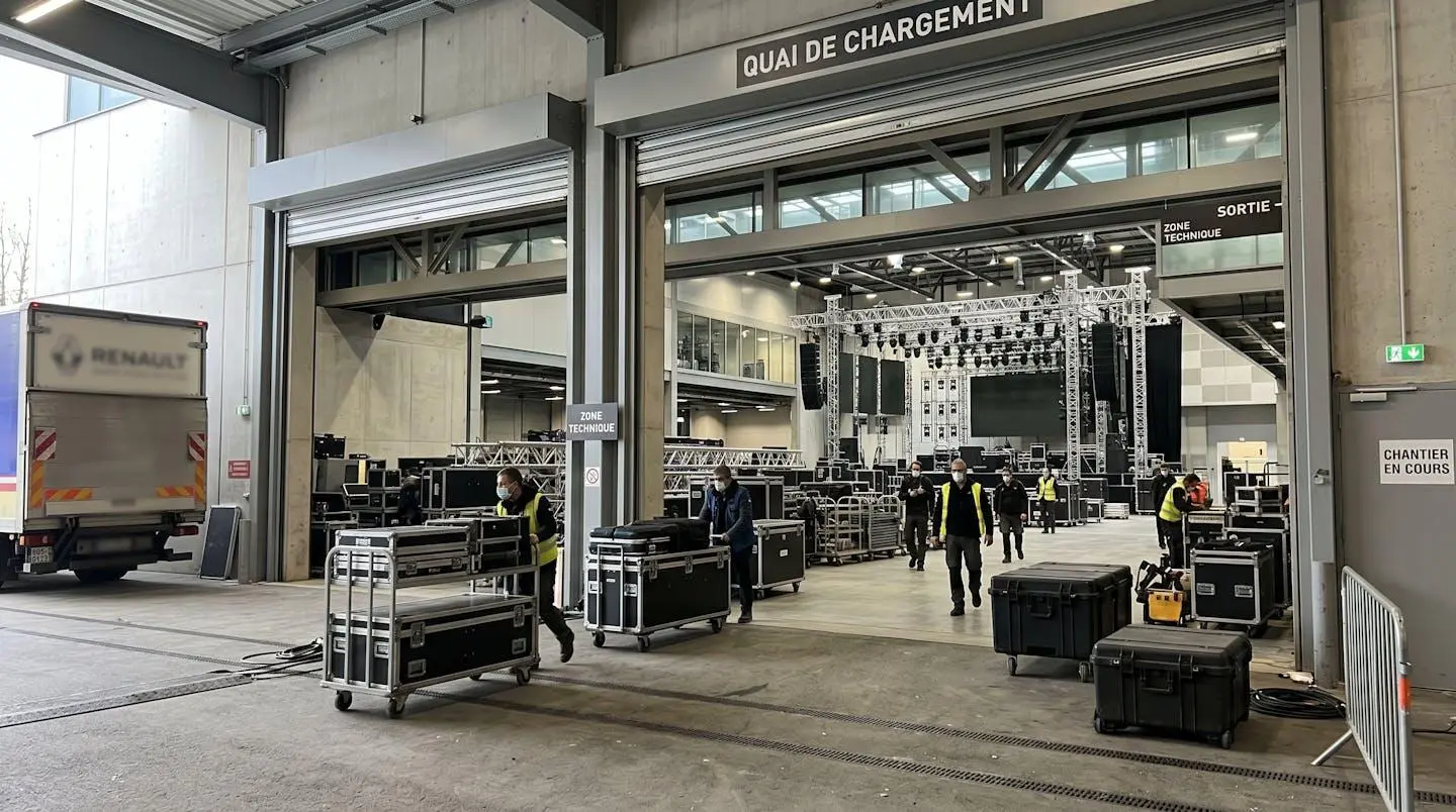 Zone de livraison matériel événementiel avec portes larges ouvertes et chariots, structure scénique visible en arrière-plan d'une salle moderne