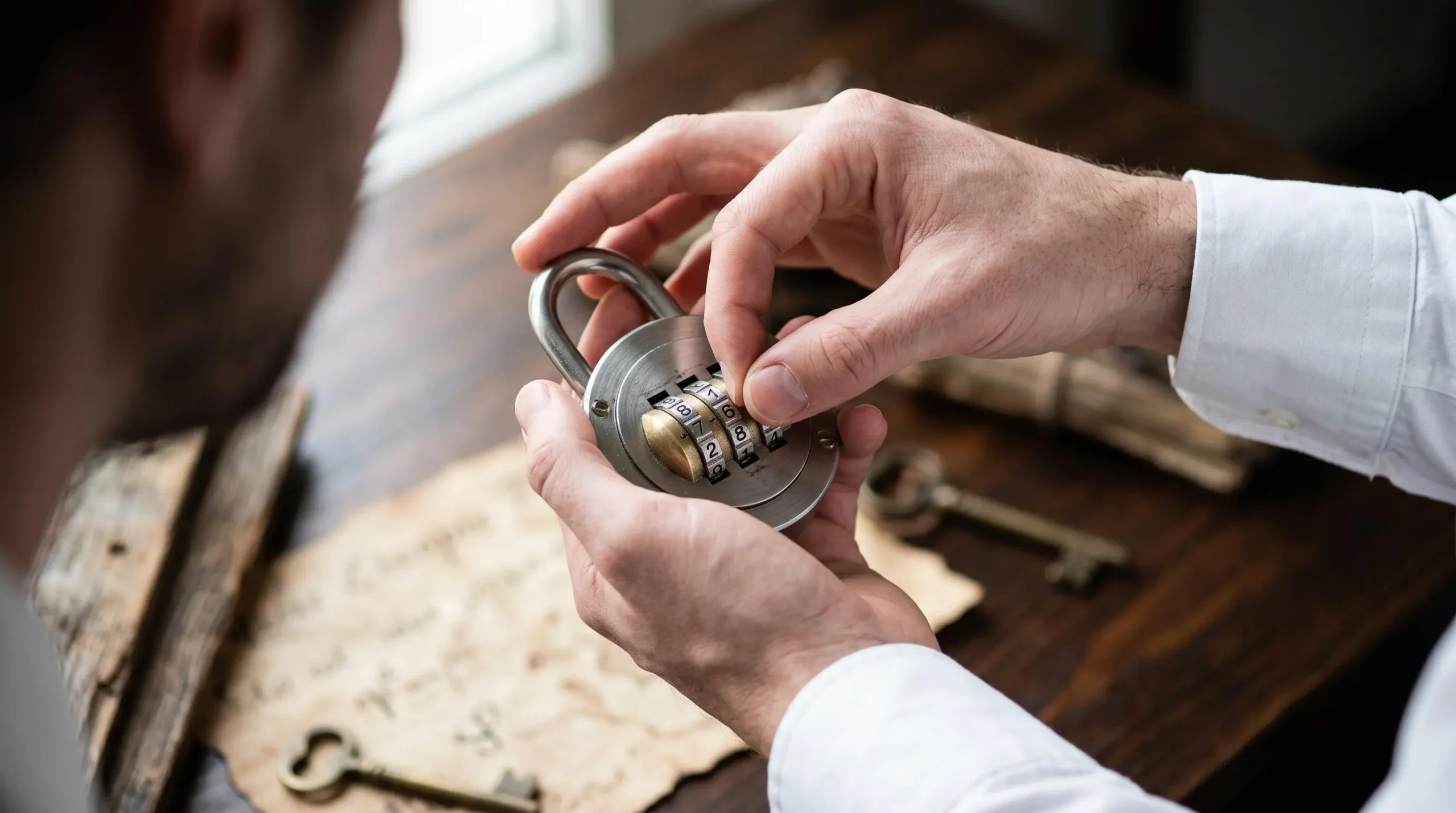 Gros plan sur les mains d'un professionnel manipulant avec précision un cadenas métallique à combinaison circulaire, doigts en action sur les molettes chiffrées, arrière-plan flouté montrant table en bois et indices d'escape game